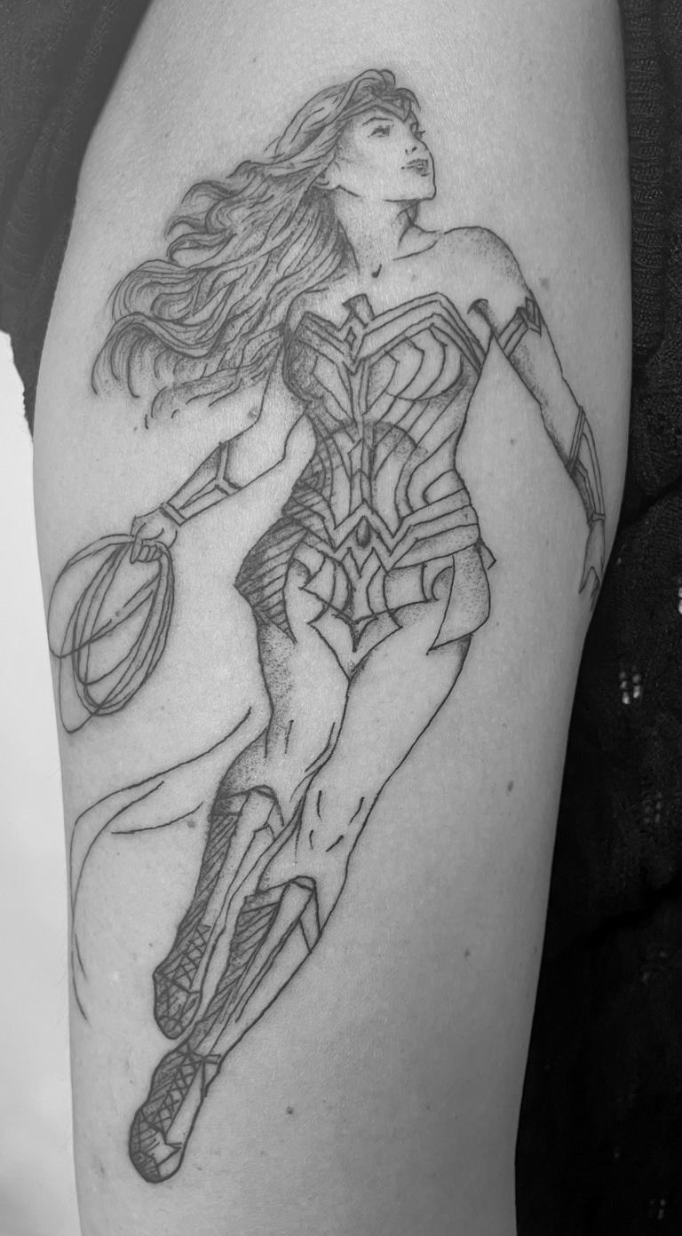 Wonder Woman in dynamischer Pose als fein gezeichnetes Tattoo auf einem Oberarm.