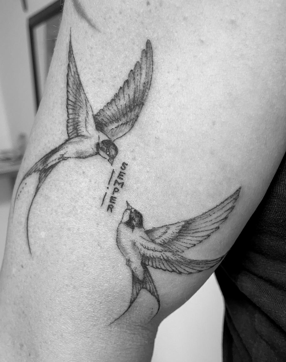 Zwei fein gezeichnete Schwalben im Flug als Tattoo auf einem Oberarm.